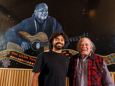 Thomas Scanlan, a young Indigenous man, with Kev Carmody, an older Indigenous man. They are standing in front of a large mural of Kev playing guitar.