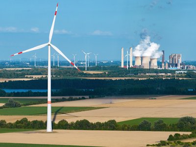 A wind turbine in a dry field with a power plant emitting steam in the background