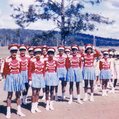 A group of teenage girls next to a smaller group of young girls. The teenagers are wearing pale blue short pleated skirts, red hats, red shirts with tassels, mid-length white socks, and white shoes. The younger children are wearing white dresses, hats, socks and shoes.